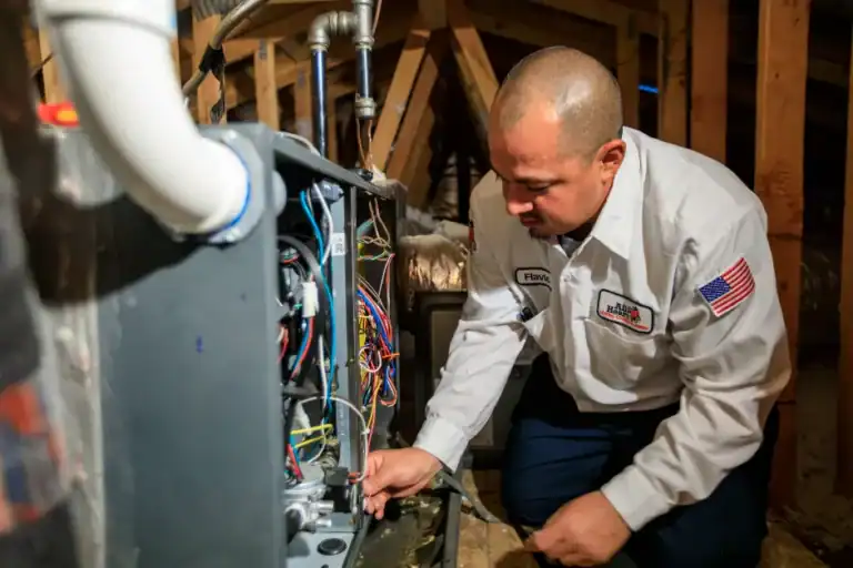 A technician in a uniform with an American flag patch is repairing or inspecting wiring inside a furnace or HVAC unit in an attic.