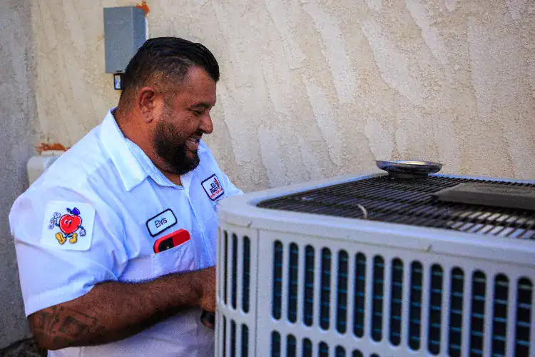 A technician in a white uniform with name tag “Elvis” works on an outdoor air conditioning unit next to a stucco wall.