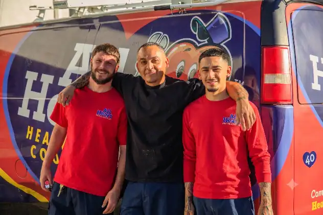 Three men stand side by side smiling in front of a red and blue company van with “A+ Heating and Cooling” written on it. Two men wear matching red shirts with the company logo.